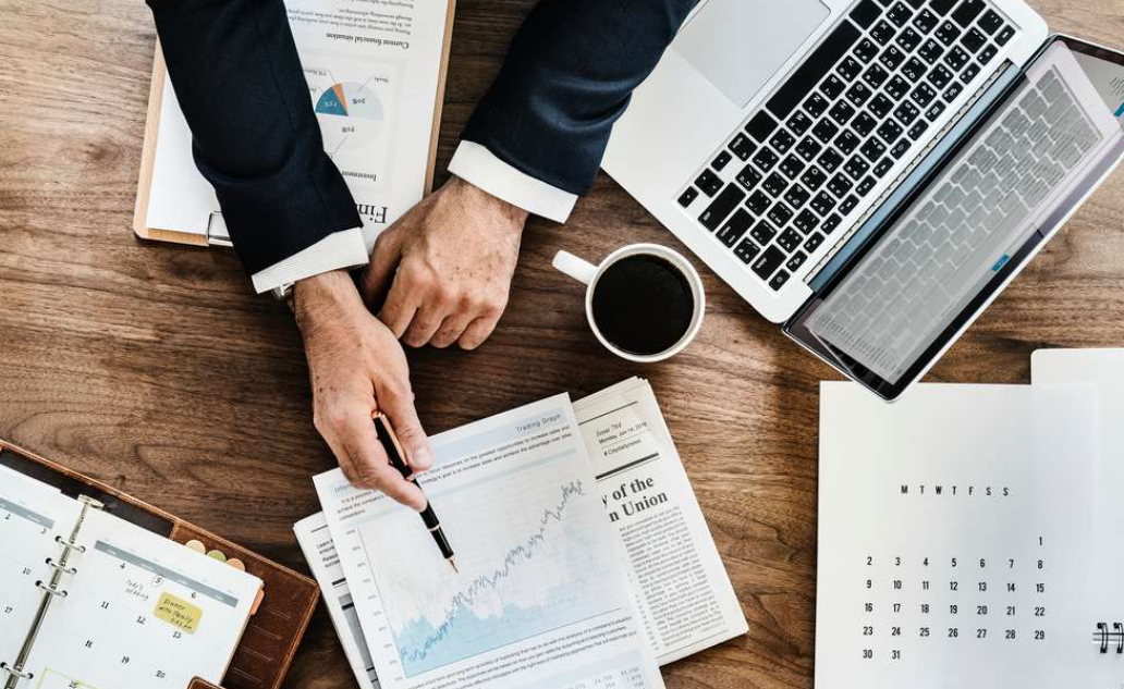 Arms outstretched over a table covered in financial reports, calendars, and a laptop