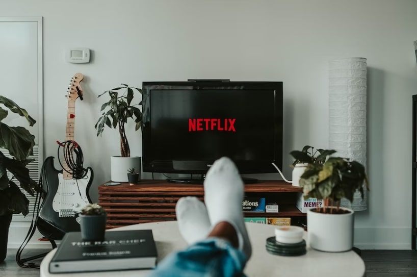 a living room with a man's feet up on a coffee table in the foreground and a TV showing the Netflix home page in the background